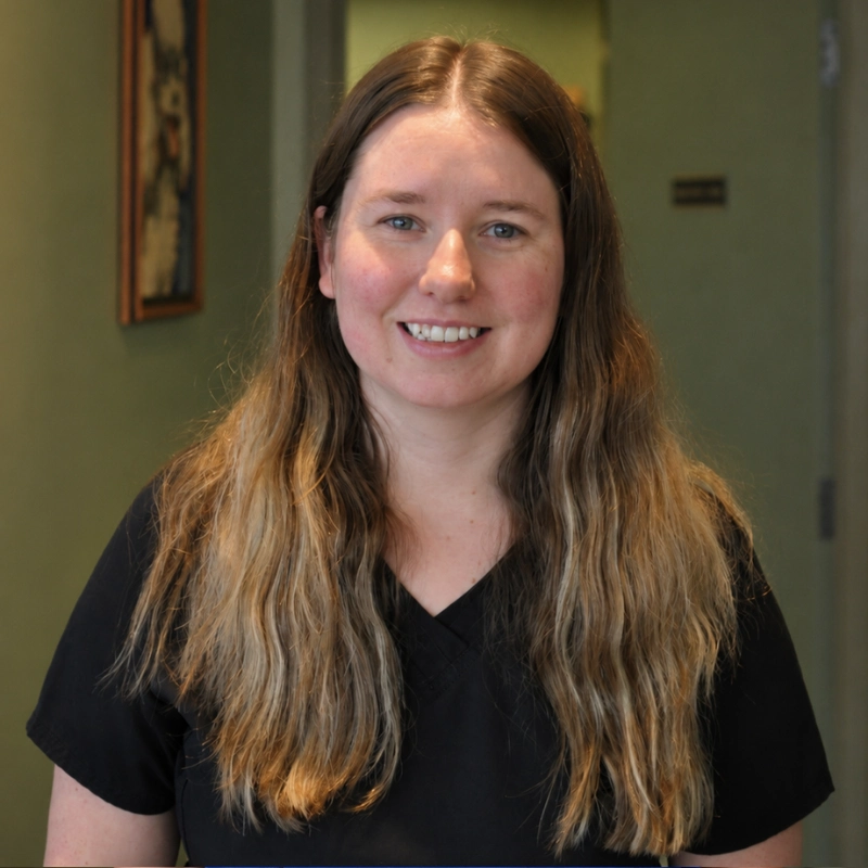 Smiling woman with long brown hair in a dark top, photographed indoors with a green wall and framed artwork in the background.