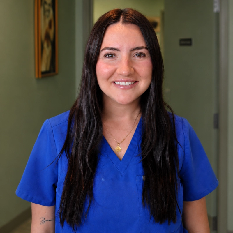 A smiling person with long dark hair wearing blue scrubs and a small gold necklace, photographed indoors in a hallway with framed artwork and neutral walls behind them.
